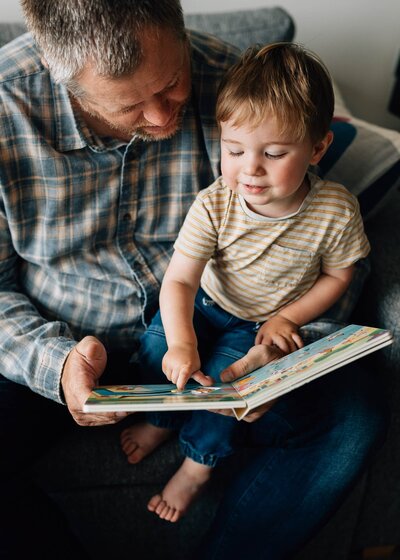 in home family photoshoot with toddler son sitting on dad's knee with book