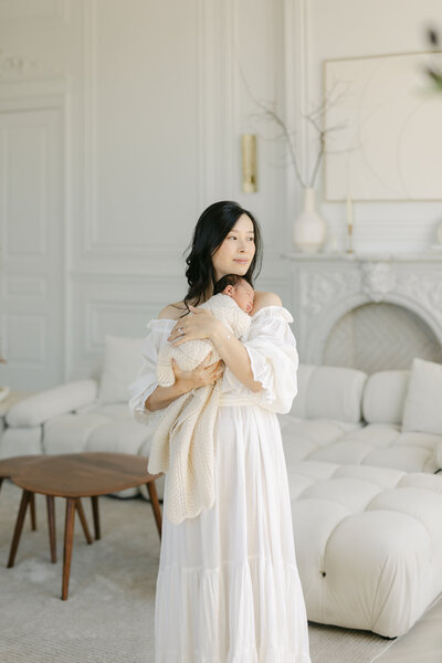 A Toronto Newborn Photographer image of a mother in a cream dress sitting on a couch snuggling her sleeping newborn baby against her chest