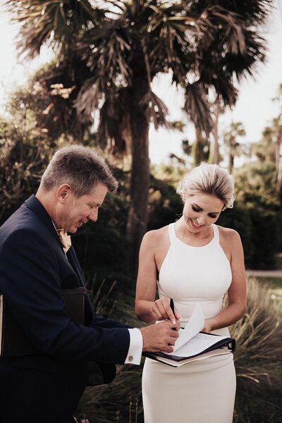 Officiant holds wedding license for bride to sign it. Palm tree in the background
