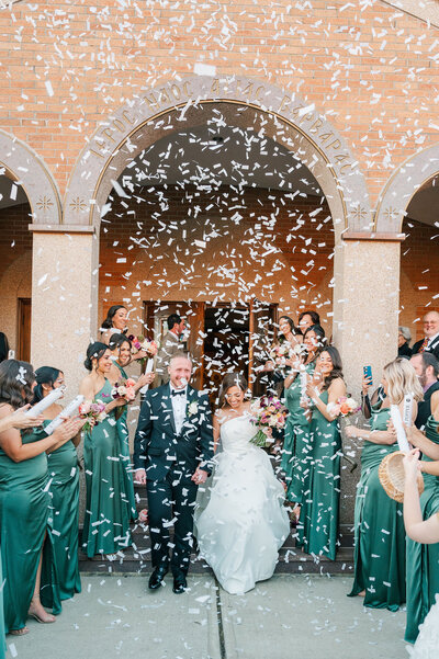 A bride and groom walk out of a Greek Orthodox church as confetti pops around them. 