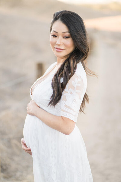 Pregnant female in white dress and wavy hair standing in a dirt path with a golden field of dry grass in the background by Orange County Maternity Photographer Melliemade Photography