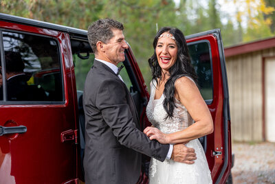 Bride and groom hugging while getting in a red Jeep Wrangler
