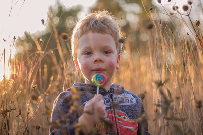 Little boy sitting on stone bench. Extremely cury brown hair. He is holding on to a stick with a roasted marshmellow on the top. Boy is smiling at camera. Child Photography