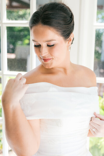 Bride with brunette hair in bun touches wedding dress at top and gazes down shoulder