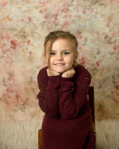 Girl sitting on a chair leaning forward on her hands. Beautiful floral wall behind her.