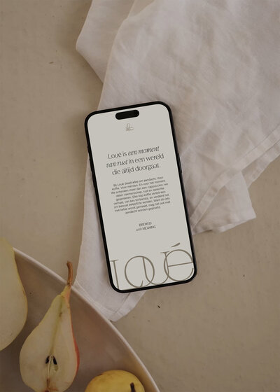 Smartphone met het merkverhaal van Loué over rust en aandacht, gepresenteerd op een beige ondergrond met peren en wit linnen servet