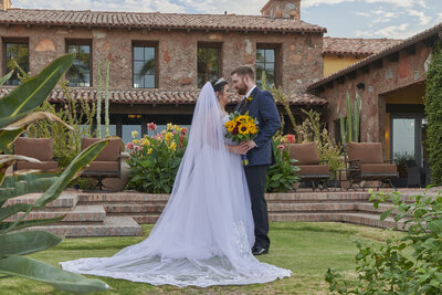 Bride and groom embrace in front of wedding venues while holding sunflower bouquet