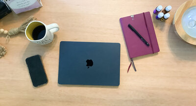 Flat lay of work essentials including coffee mug, MacBook, essential oils, phone, and journal on a wooden desk