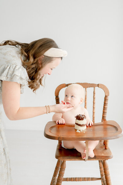 a baby smiles at his mom as she feeds him a bite of smash cake