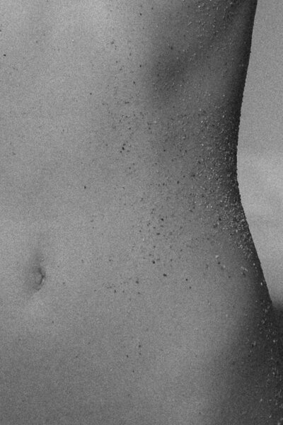 Close-up black and white photograph of a woman's torso with fine grains of sand scattered across the skin, capturing natural texture and sensual detail.