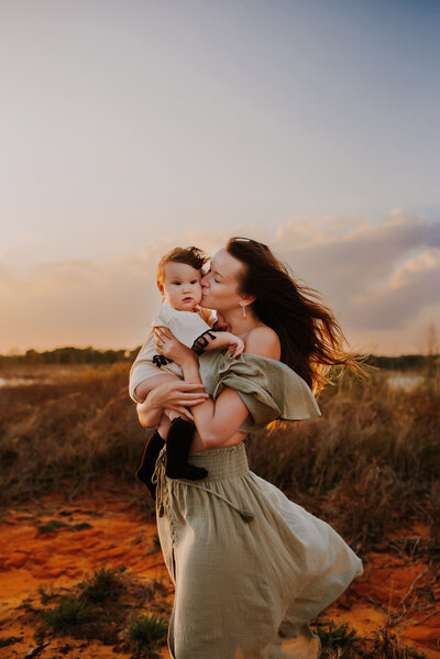 A mom holding her baby girl in her arms while kissing her cheek at Murrell park in Flower Mound, Texas