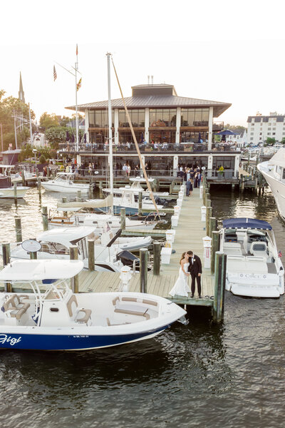 A couple stands at the end of a dock surrounded by boats, with a waterfront restaurant in the background. The setting sun casts a warm glow on the scene, highlighting the boats and bustling venue.