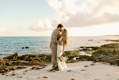 Bride and groom kiss on rocky Cancun shoreline with turquoise waves and golden sunlight behind them.