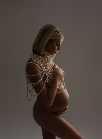 Studio image of a pregnant woman wearing layered pearl body jewelry in a soft spotlight, highlighting high-end maternity accessories and styling options