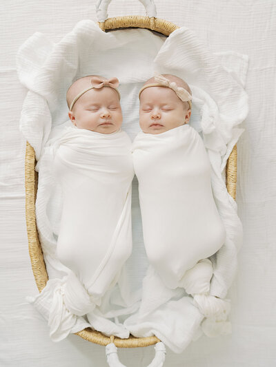 Two swaddled newborn twin girls are peacefully sleeping in a basket, nestled closely together. Their tiny, delicate features are visible as they rest soundly, wrapped in soft blankets, creating an adorable and serene moment.