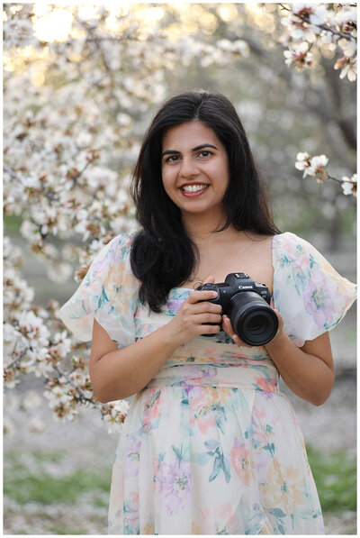 Female photographer holding a professional camera and gazing directly at viewer while posing in almond orchard with white blossoms during sunset