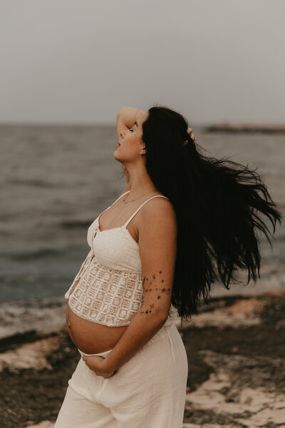 A couple embraces in a golden field during a maternity photoshoot at sunset, with the expectant mother wearing a rust-colored dress and the partner in a white shirt and teal pants, surrounded by tall grass and a scenic natural landscape in Peapack, NJ.
