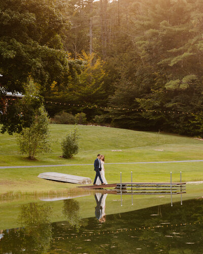 A picturesque scene of the bride and groom walking out onto the dock during their wedding at Juniper Springs Wedding Barn in Corinth, NY.