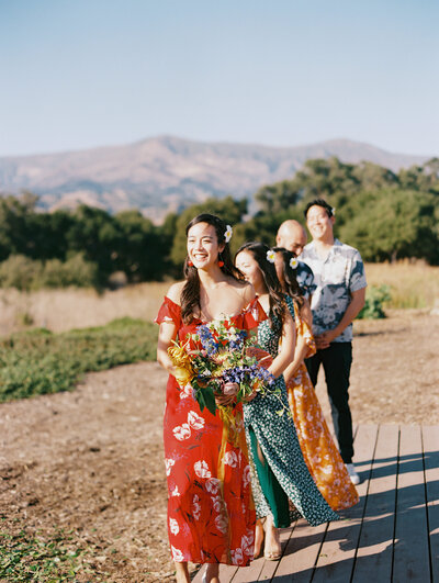 The wedding party smiles while looking at the couple marrying in the scenic California Coast, with the maid of honor holding the bridal bouquet designed by Moonlight Floral Co, by photographer My Sun and Stars Co.