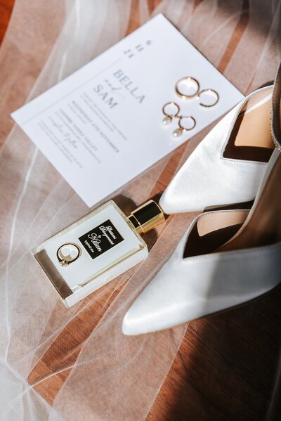 White wedding shoes with a white wedding invitation, wedding rings, and perfume