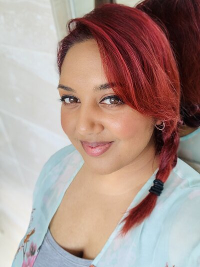 Image shows a head and shoulders portrait photo of Life Alignment Coach Naaila with her red hair braided to the side, with a light blue sheer top and looking at the camera.
