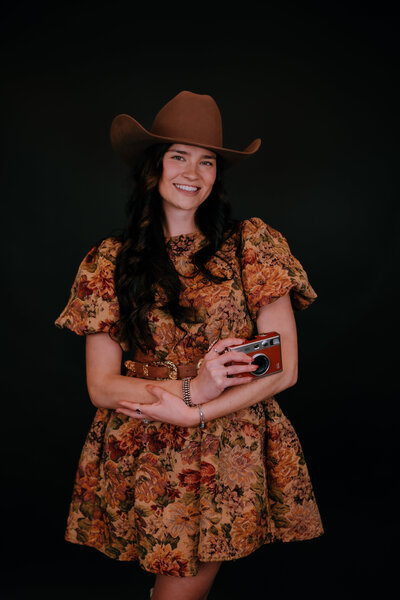 woman holding a camera while smiling and wearing a dress and cowgirl hat