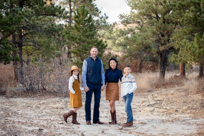 A family of 4 stand together holding hands in a forest trail at sunset