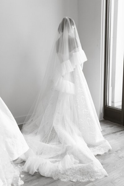 Bride in an off-shoulder lace gown with a long veil standing near a window.