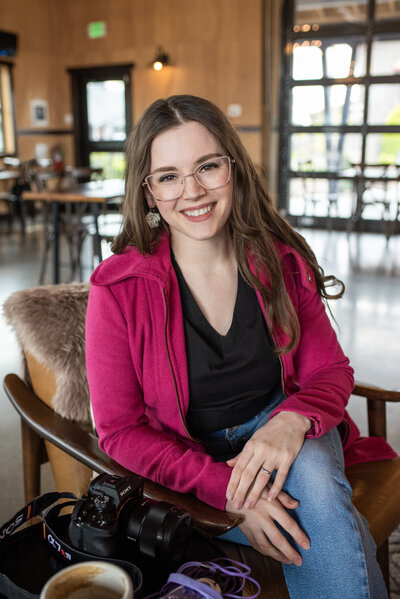 Professional photographer Lana Stewart sitting for headshot wearing magenta coat and jeans at Fika cafe