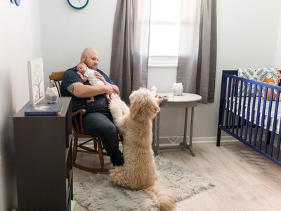 dad holding newborn baby boy with family dog in nursery