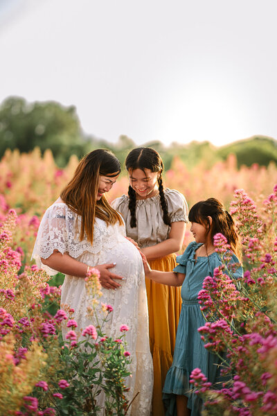 mom in white maternity dress in a field of wildflowers with her 2 daughters