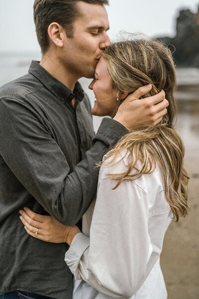 A man kisses a woman's forehead as they embrace on the Oregon coast. 