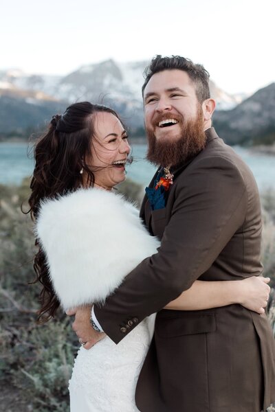 A bride and groom laugh together while in front of an alpine lake on their mammoth wedding day. 