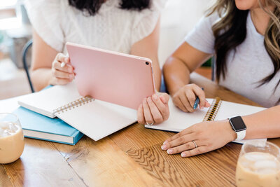 Two women strategizing at a table over a notebook and iPad.