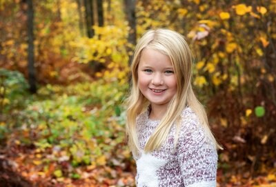 A young pre-teen girl with blonde hair and blue eyes smiling with the fall colors surrounding her outside at Studio 64 Photography.