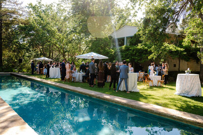 Elegant BP Collective poolside event with guests mingling under trees, cocktail tables with white umbrellas, and a serene garden setting.