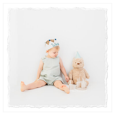 The photo shows an adorable two-year-old boy celebrating his birthday with a portrait session. He is dressed in a stylish teal romper and wears a birthday crown adorned with his name. A small stuffed bear and a wooden birthday cake add to the festive atmosphere. The photograph is taken against an all white background, creating a pure and clean look. It is beautifully captured by photographer Bri Sullivan.