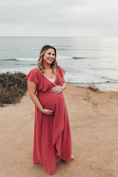 a pregnant woman in a pink dress holds her baby bump while walking at Sunset Cliffs in San Diego