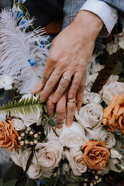 A close-up of a couple's hands with matching gold wedding bands resting on a bouquet of white and burnt orange roses, accented with greenery, blue flowers, and delicate feathers, symbolizing love and unity.