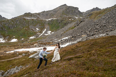A couple hikes hand in hand through rocky alpine terrain with patches of snow during their adventurous Alaska mountain elopement, captured by Sydney Breann Photography.