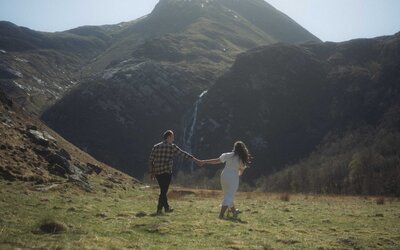 Couple strolling near Steall Waterfall, sharing a loving glance amidst the breathtaking scenery of the Scottish Highlands, full of romance and adventure