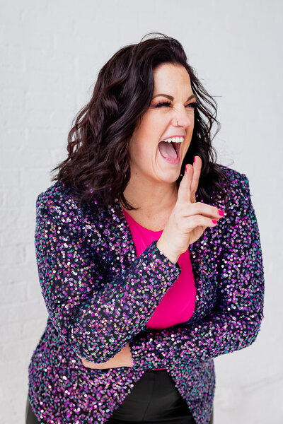Jessi Cabanin laughing and giving a peace sign with her fingers while wearing a purple sequin jacket and hot pink shirt.