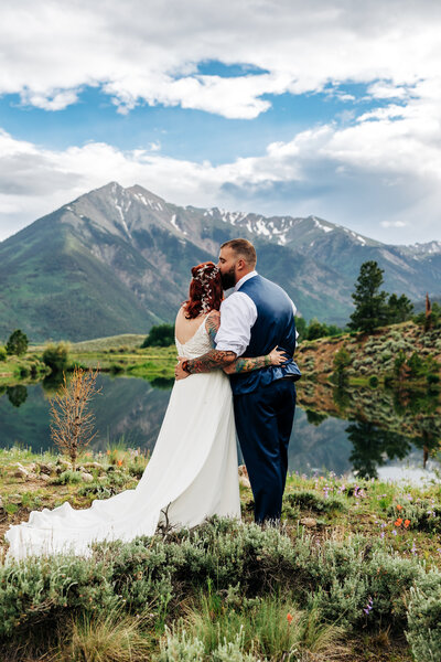 couple is facing away and holding each other. the groom is kissing the bride on the head. There is a big mountain in the background with a lake and greenery 