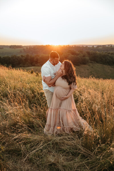 A pregnant mother wearing a gorgeous gown holding her belly while her husband holds her chin up to gaze at him. 