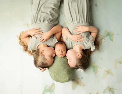 Three children lie side by side; two sisters in gray dresses hug a sleeping baby in green. Soft pastel butterflies decorate the serene background.