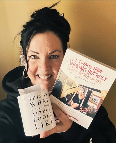 Barb Drummond smiling and holding her book titled "I Finally Have the Smoking Hot Body I Have Always Wanted (having been cremated)" in one hand and a white mug in the other that reads "This is what a published author looks like."