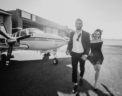 Black and white photo of a man and women, holding hands. There are on a airport runway walking away from a small plane.