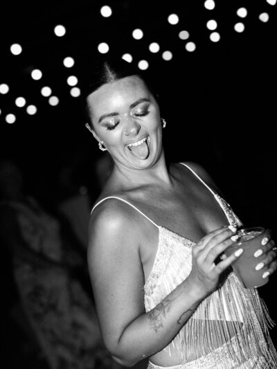 bride wearing a sequin dress with a drink in her hand sticks her tongue out on the dance floor