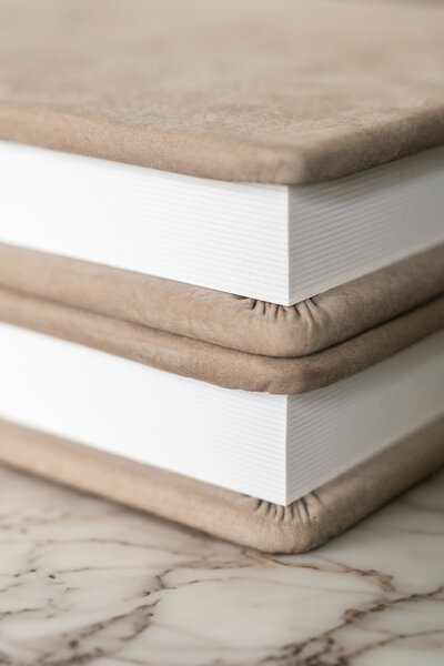 Close-up of two beige hardcover books stacked on a marble surface, showcasing the edges of the covers and pages.