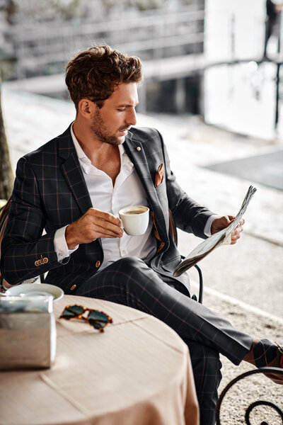 A man in a navy suit and unbuttoned white shirt sips espresso, reading the paper—waiting, knowing he’ll steal her breath away.